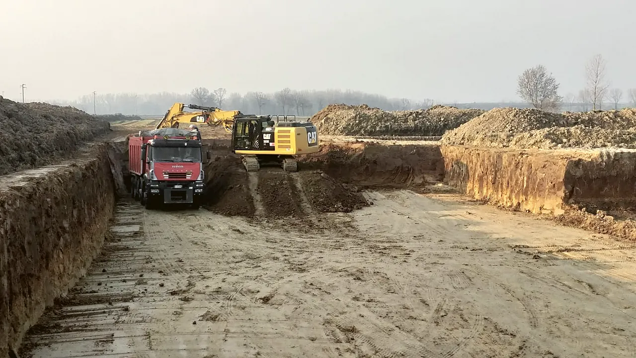 Cava in coltivazione: escavatore Caterpillar sul fronte, camion in caricamento e piste di servizio fra i cumuli