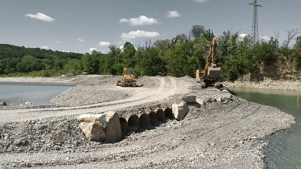 Intervento di regimazione fluviale: escavatore e apripista al lavoro sulla realizzazione di un attraversamento con tubazioni in un alveo